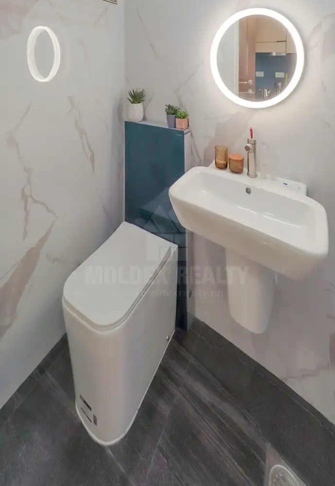 Studio bathroom shown with a wall basin, round lit mirror and toilet